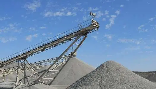 Industrial conveyor belt system depositing gravel onto a large pile at an outdoor processing plant. The sky is bright blue with scattered clouds, and the background shows additional gravel piles and flat terrain.